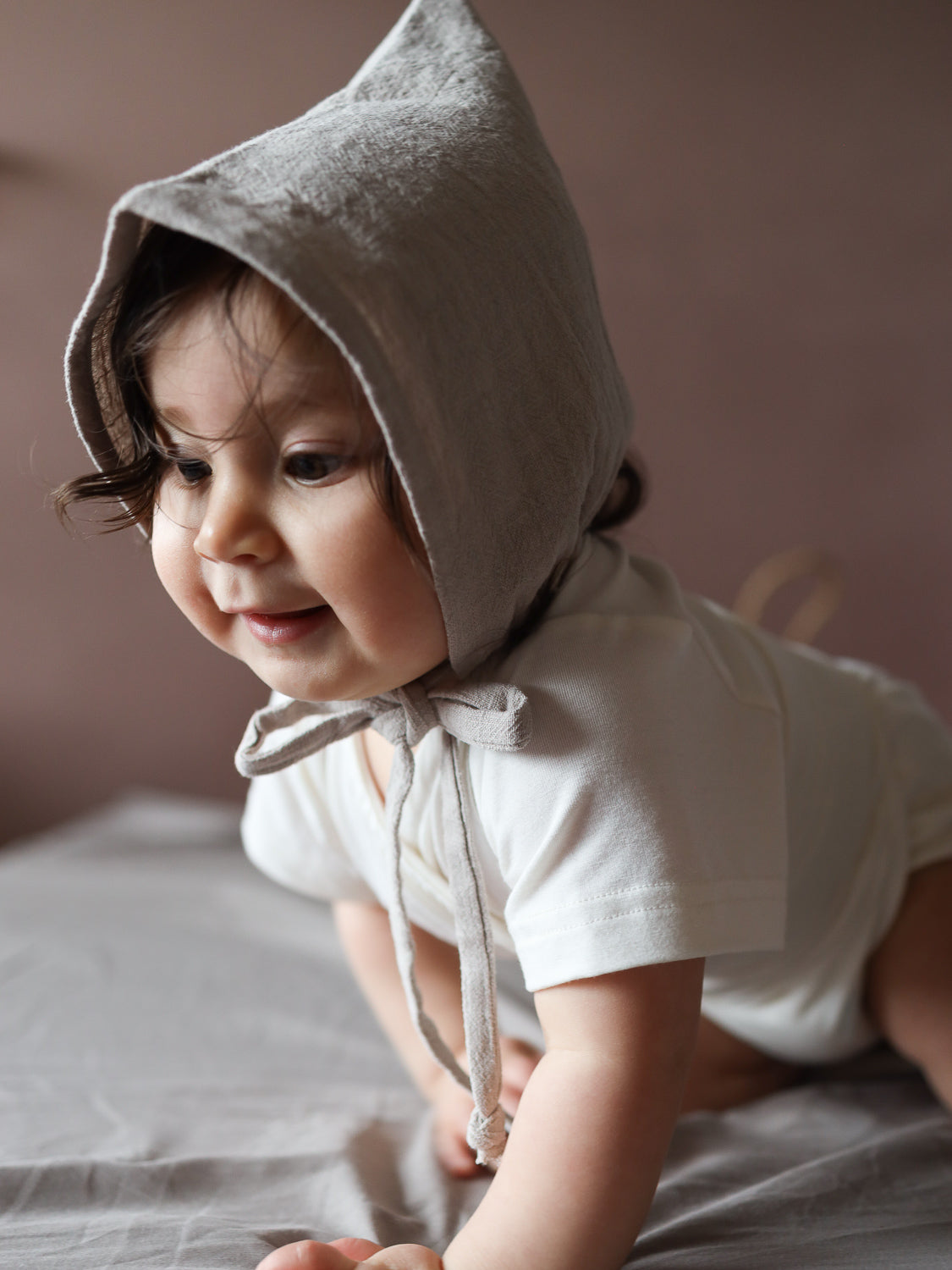 Kleines Kind in einem schlichten, cremefarbenen Onesie, trägt eine Zwergenmütze Leinen - Sand. Die weiche, neutrale Mütze schützt Kopf und Ohren, ideal für jede Jahreszeit.