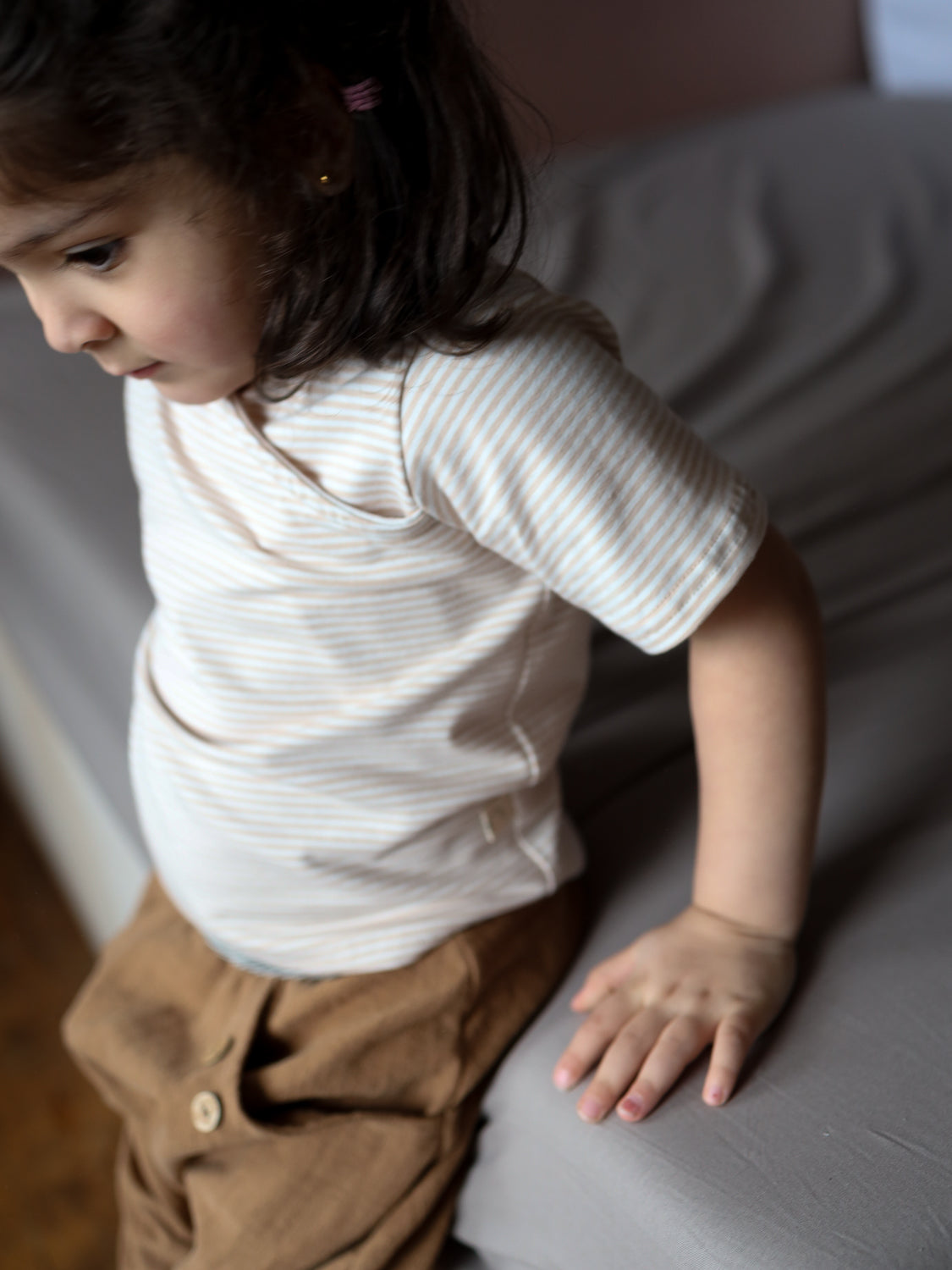 Ein Kleinkind trägt das Kurzes Wickelshirt Baumwolle - Ringel Sand, mit feinen Streifen und Wickelausschnitt, sitzt am Bettrand, kombiniert mit lockeren braunen Hosen, in einem sanft beleuchteten Schlafzimmer.