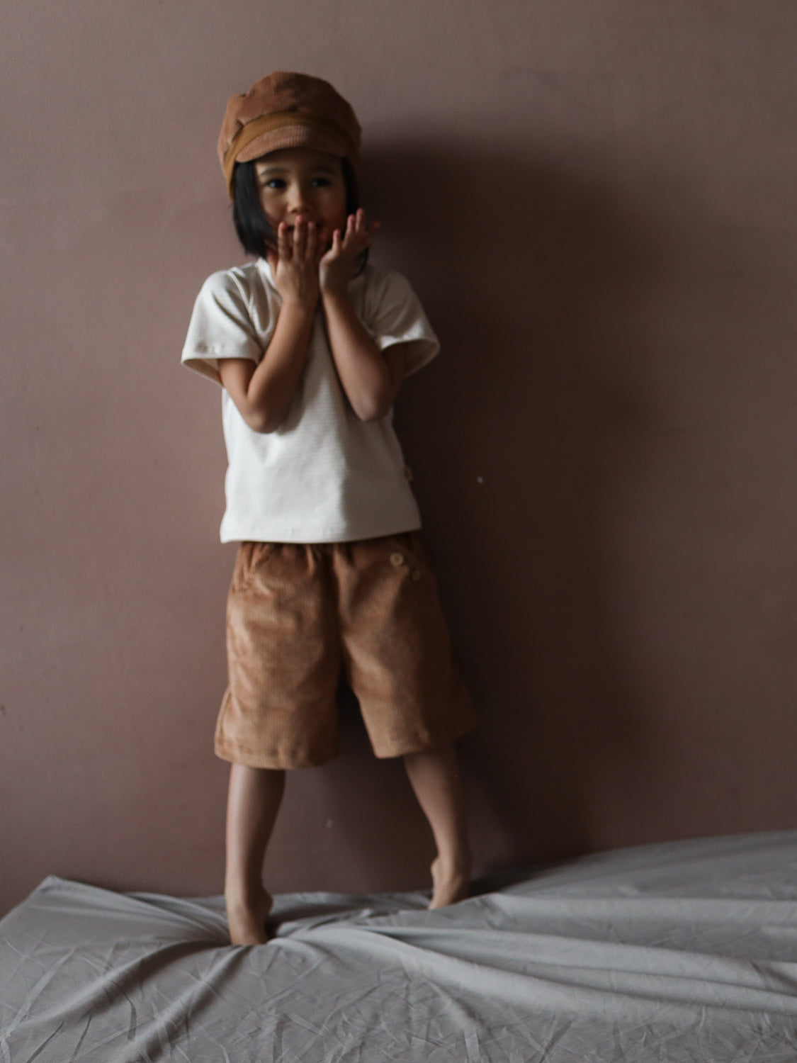 Ein Kind trägt das Kurzes Wickelshirt Baumwolle - Ringel Sand, ein weißes kurzärmeliges Oberteil, vor einer braunen Wand. Es steht barfuß auf einem grauen Laken, die Hände nah am Gesicht.