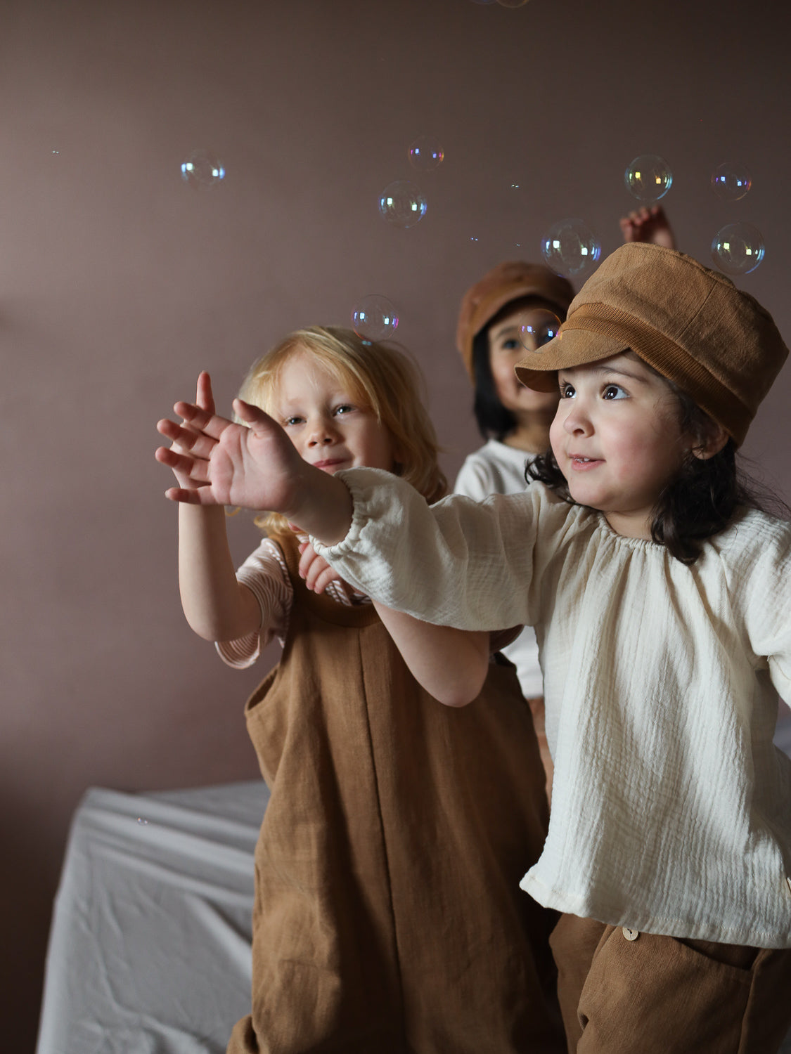 Drei Kinder spielen mit Seifenblasen. Im Vordergrund trägt ein Kind die Michelmütze Leinen - Weizen, eine kordähnliche Kappe, während es freudig die Hände zu den Seifenblasen ausstreckt.