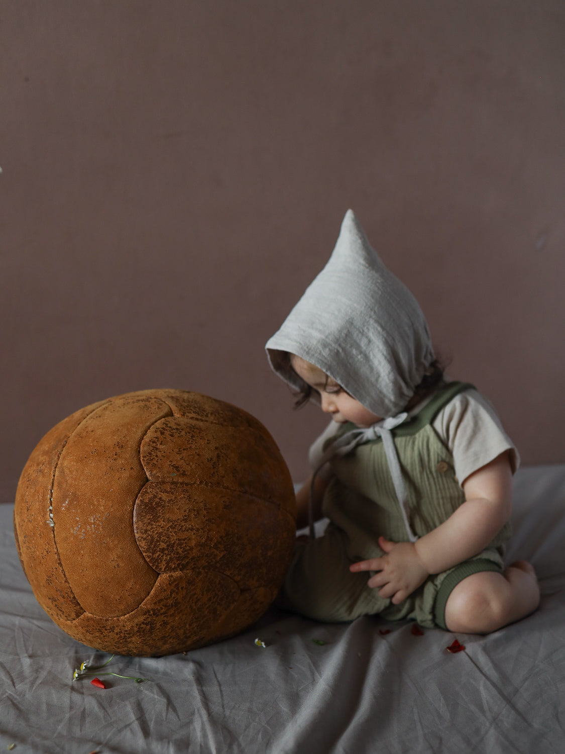 Kleinkind in Baumwoll-Strampler und Zwergenmütze Leinen - Sand sitzt vor großem, vintage-braunem Lederpouf auf Stoffuntergrund, umgeben von verstreuten Blumenblättern.