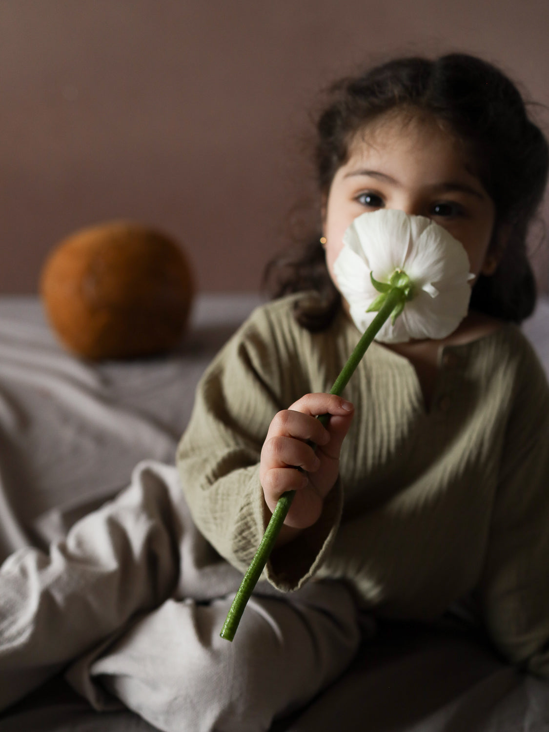 Kind mit dunklem, welligem Haar trägt Hemdchen Musselin - Salbei, locker geschnitten, auf neutralen Stoffen sitzend, hält eine weiße Blume, weiche, atmungsaktive Textur für warme Tage.