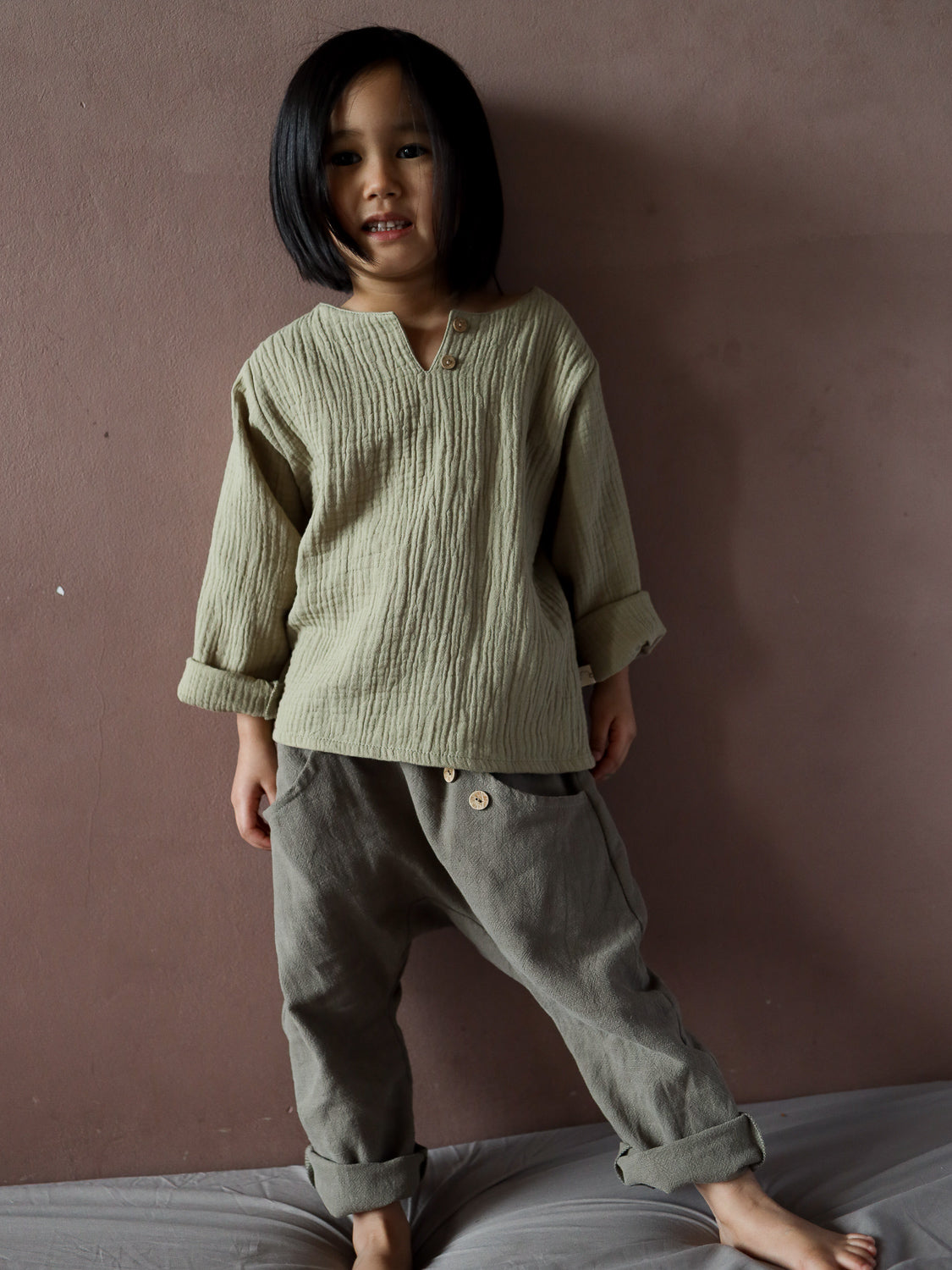 Ein Kind trägt das Hemdchen Musselin - Salbei, ein locker sitzendes, langärmliges Oberteil mit kleinem Knopfkragen, aus weichem, atmungsaktivem Bio-Musselin, ideal für warme Tage.