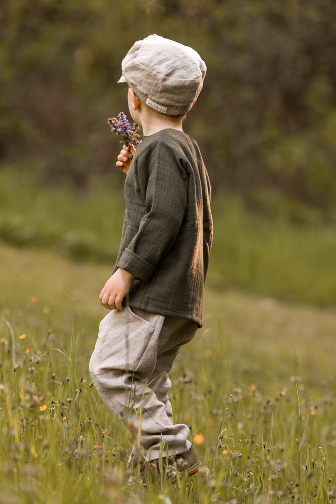 Ein Kind steht in einem Wiesenfeld und hält lila Wildblumen. Es trägt die Michelmütze Leinen - Sand, eine weiche, neutrale Mütze, passend zu heller Leinenhose und dunklem Oberteil.