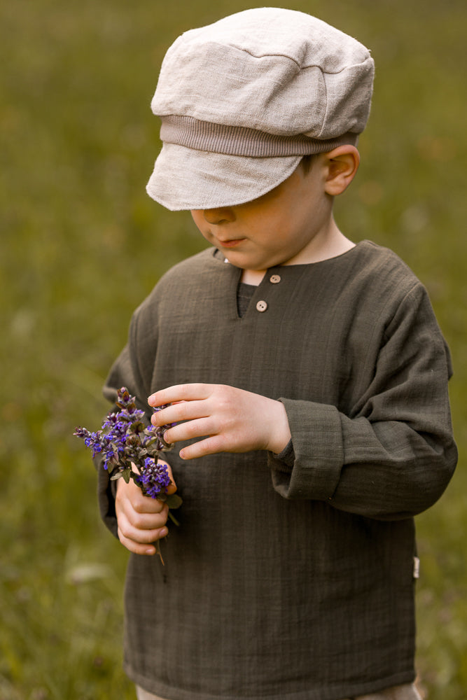 Ein Junge trägt die Michelmütze Leinen - Sand, eine weiche, beige Kappe, während er lila Wildblumen betrachtet. Sein olivgrünes Langarmshirt ergänzt die natürliche, ruhige Umgebung.