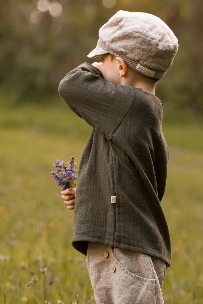 Ein Kind trägt die Michelmütze Leinen - Sand, eine weiche, neutrale Kappe, in einem natürlichen Setting, ergänzt durch lockere, erdfarbene Kleidung und einen Strauß lila Wildblumen.