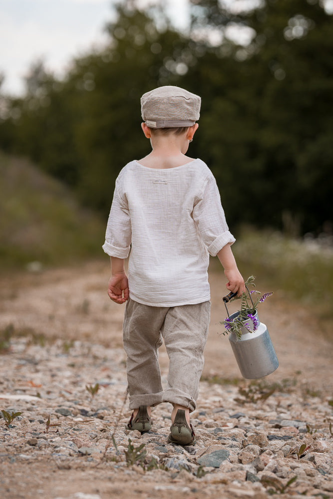 Ein Kind trägt die Michelmütze Leinen - Sand, während es auf einem Kiesweg läuft. Es hält eine kleine Metallkanne mit lila Wildblumen, umgeben von Natur und Bäumen im Hintergrund.