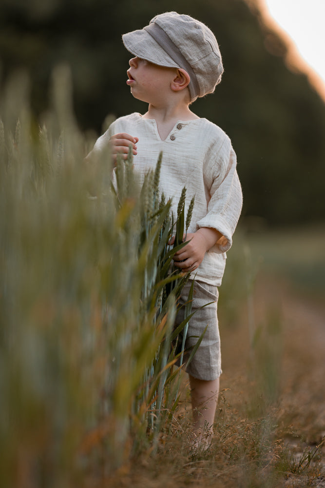 Ein Kleinkind steht am Rand eines Getreidefelds und trägt die Michelmütze Leinen - Sand. Die weiche, neutrale Kappe spendet Schatten, während er die Pflanzen sanft mit der Hand berührt.