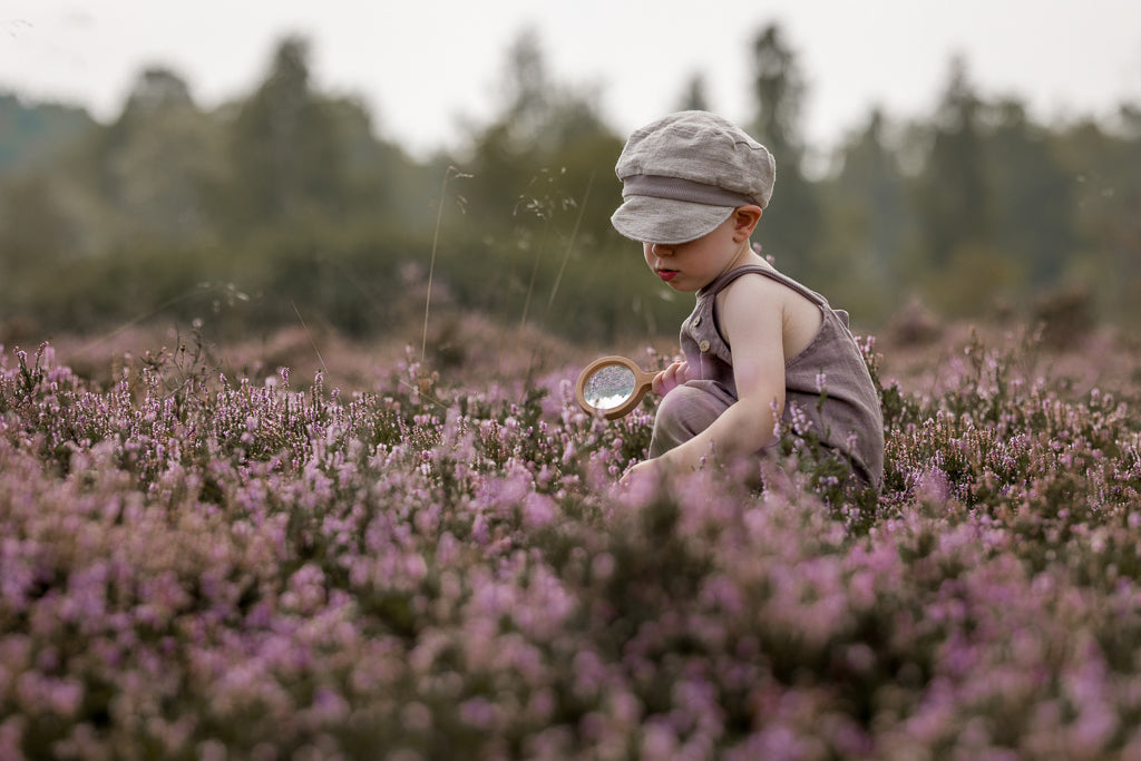 Ein Kleinkind im Feld untersucht Blumen mit einer Lupe, trägt die Michelmütze Leinen - Sand, eine hellgraue Schiebermütze, und einen mauvefarbenen Strampler.