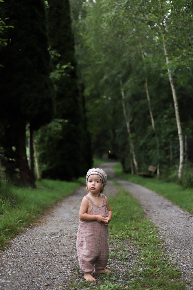 Ein Kleinkind trägt die Kurze Schlupflatzhose Bio-Musselin - Puder, während es barfuß auf einem schmalen Waldweg steht, umgeben von hohen Bäumen.