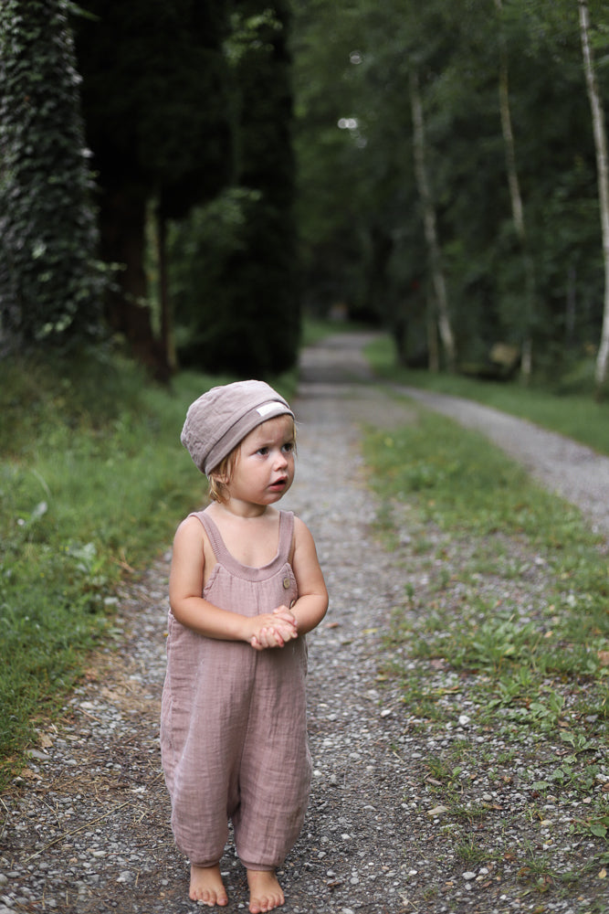 Ein Kleinkind trägt die Kurze Schlupflatzhose Bio-Musselin - Puder, steht barfuß auf einem Kiesweg im Grünen. Der ärmellose Einteiler bietet Bewegungsfreiheit in natürlicher Umgebung.