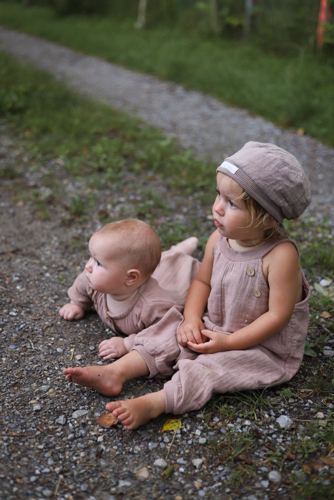 Zwei Kleinkinder tragen die Kurze Schlupflatzhose Bio-Musselin - Puder, sitzen auf einem Kiesweg. Die luftigen Einteiler sind bequem und handgenäht, ideal für Sommerabenteuer.