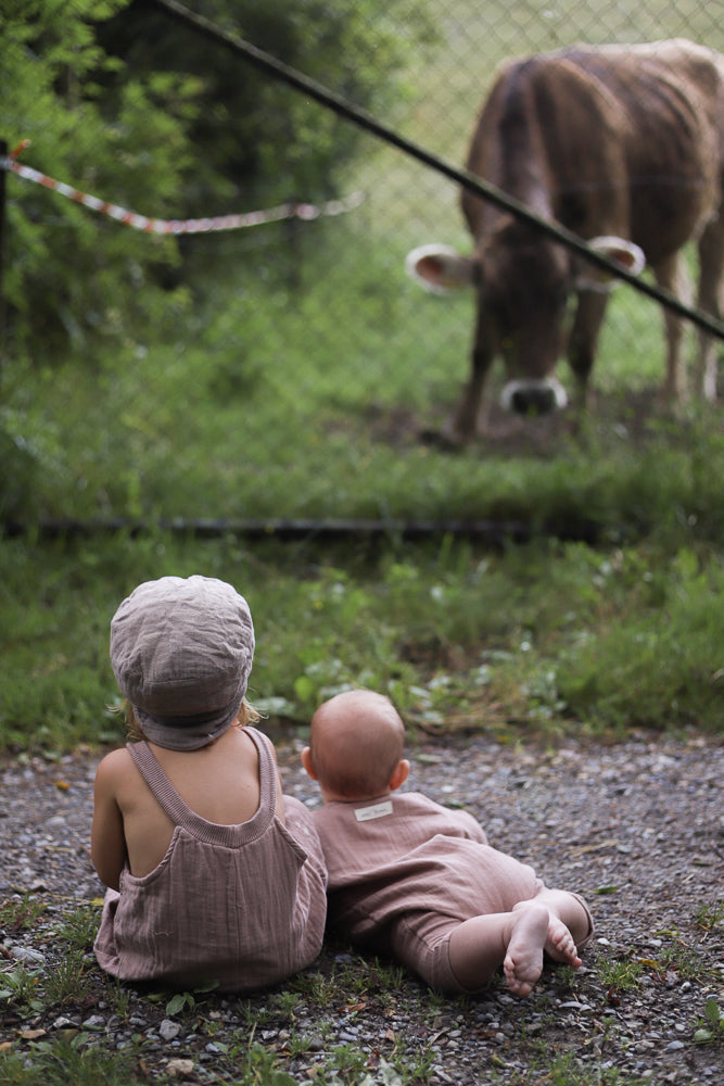 Zwei Kleinkinder, eines trägt die Kurze Schlupflatzhose Bio-Musselin - Puder, sitzen auf Kies und schauen durch einen Zaun eine Kuh an. Der Romper ist locker und bequem.
