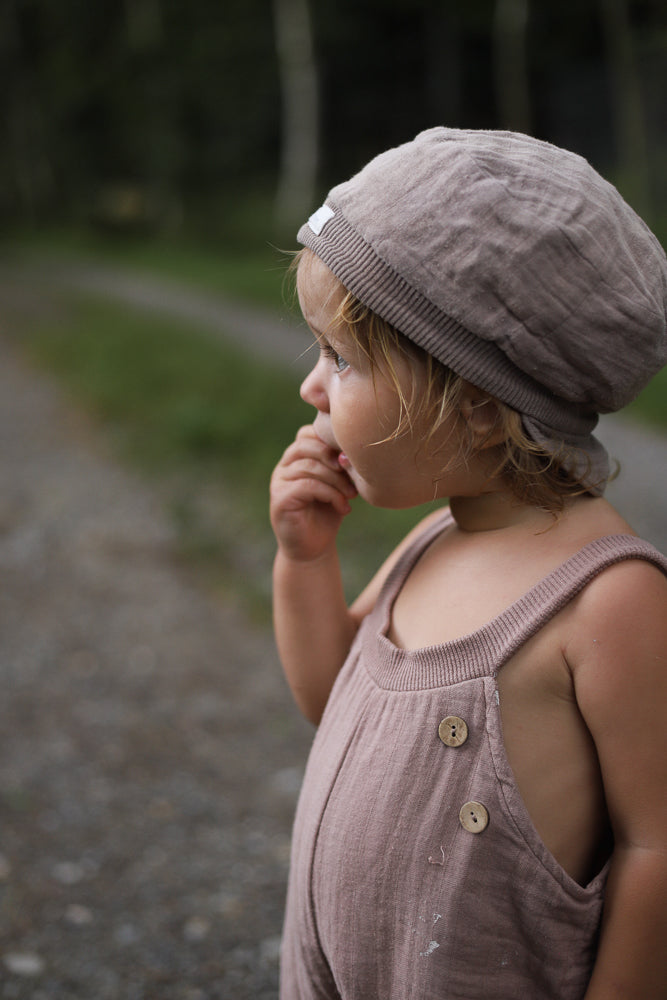 Kleinkind auf einem Kiesweg trägt die Kurze Schlupflatzhose Bio-Musselin - Puder, einen locker sitzenden Leinen-Einteiler mit Holzknöpfen, handgenäht in Berlin, ideal für sommerlichen Komfort und Bewegungsfreiheit.