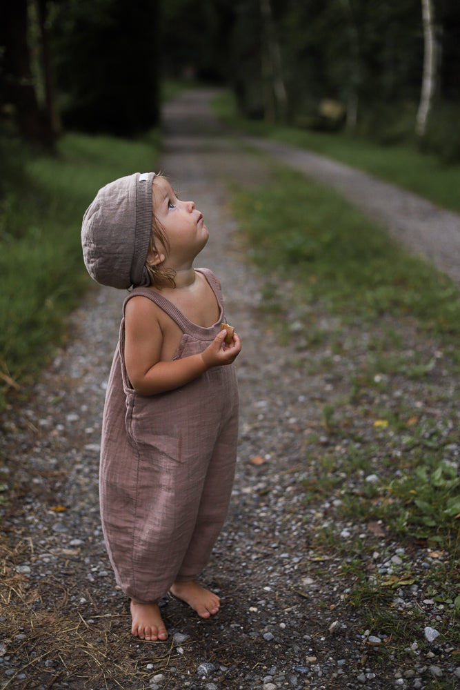 Kind in Kurze Schlupflatzhose Bio-Musselin - Puder steht barfuß auf einem Waldweg, hält einen Snack und schaut nach oben. Die bequeme, handgenähte Latzhose bietet Bewegungsfreiheit für sommerliche Abenteuer.