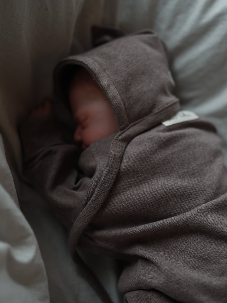 Ein schlafendes Neugeborenes in einer weichen, braunen Zwergenmütze Feinstrick - Praline Meliert liegt auf einer hellen Decke. Die Mütze hat eine charakteristische Zipfelform und hält den Kopf warm.