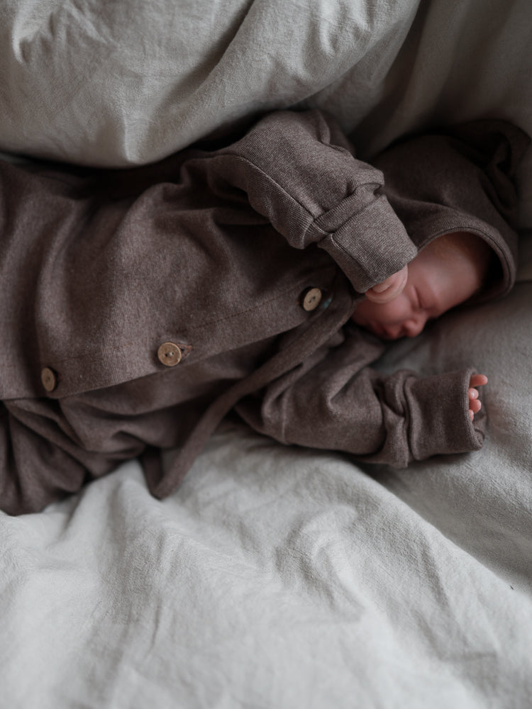 Schlafendes Baby in Zwergenmütze Feinstrick - Praline Meliert, einem weichen, braunen, kapuzenartigen Einteiler mit Holzknöpfen, auf grauer Bettwäsche liegend. Die Mütze zeigt die charakteristische Zipfelform.