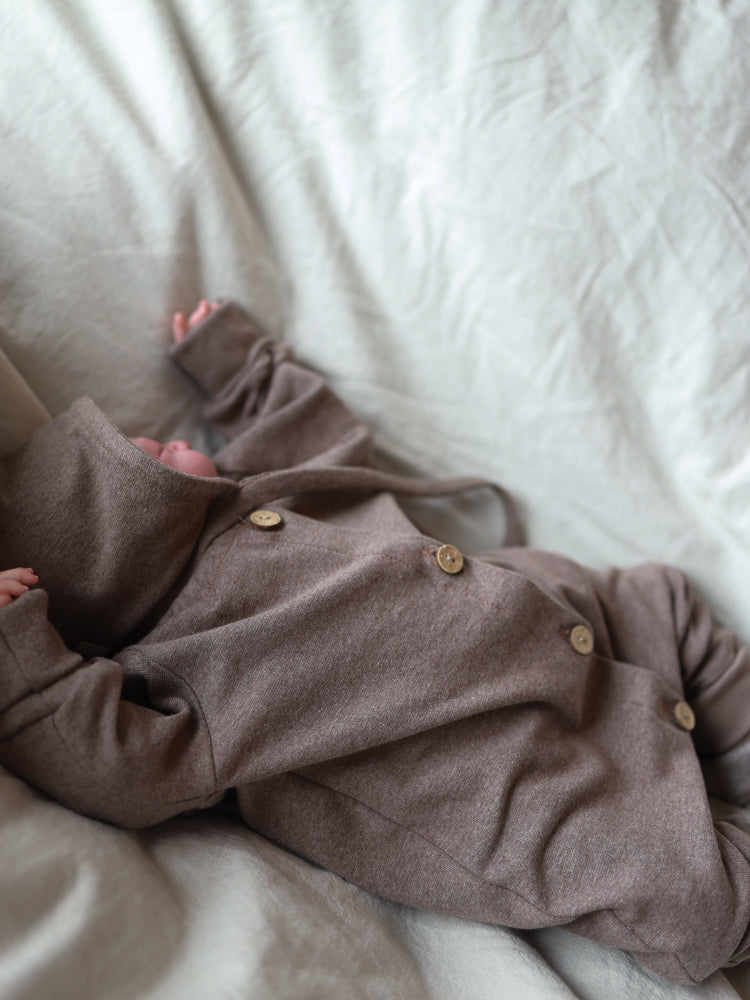 Ein Neugeborenes liegt auf einer weichen Decke, trägt einen braunen, melierten Zwergenmütze Feinstrick - Praline Meliert, mit Kapuze und Holzknöpfen, die für Wärme und Komfort sorgen.