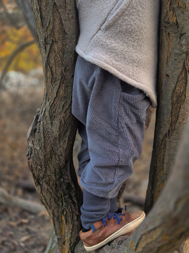Kind trägt Knickerbocker Breitcord Stretch - Maus, während es zwischen Baumstämmen steht. Die Hose aus weichem Breitcord bietet Bewegungsfreiheit, ideal für kleine Entdecker:innen bei kühlerem Wetter.