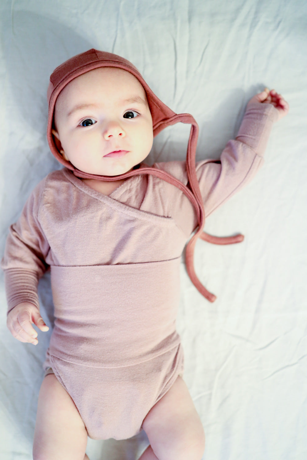 Babyhäubchen Kaschmir-Mix - Zimt: Ein Baby liegt auf einem Bett und trägt eine weiche, elastische Kaschmir-Mix-Mütze mit langen Bändern für optimalen Halt. Baby Body für Erstausstattung Baby.