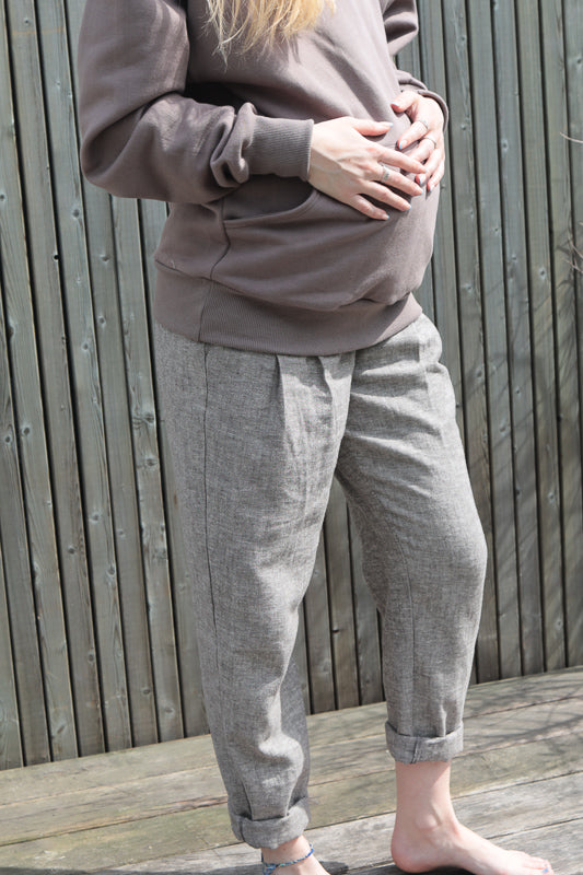 Schwangere Frau im Freien trägt Bundfaltenhose Fischgrat, legt Hände auf Bauch. Hose wirkt lässig, aus Denim, mit sichtbaren Taschen.