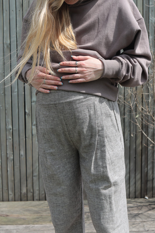 Frau steht, Hände auf dem Bauch, trägt Bundfaltenhose Fischgrat, lässiger Street-Fashion-Stil mit Fokus auf Taschen und Denim-Optik, betont durch die entspannte, alltägliche Haltung.