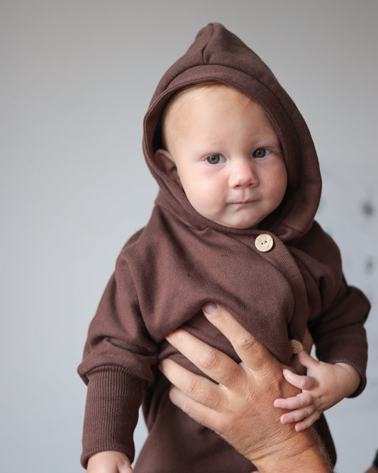 Ein Baby trägt den Kuschelanzug Bio-Sweat - Schoko mit Wichtelkapuze, ideal für Komfort und Wärme, aus weicher Bio-Baumwolle, perfekt für verschiedene Jahreszeiten und langes Tragen geeignet.