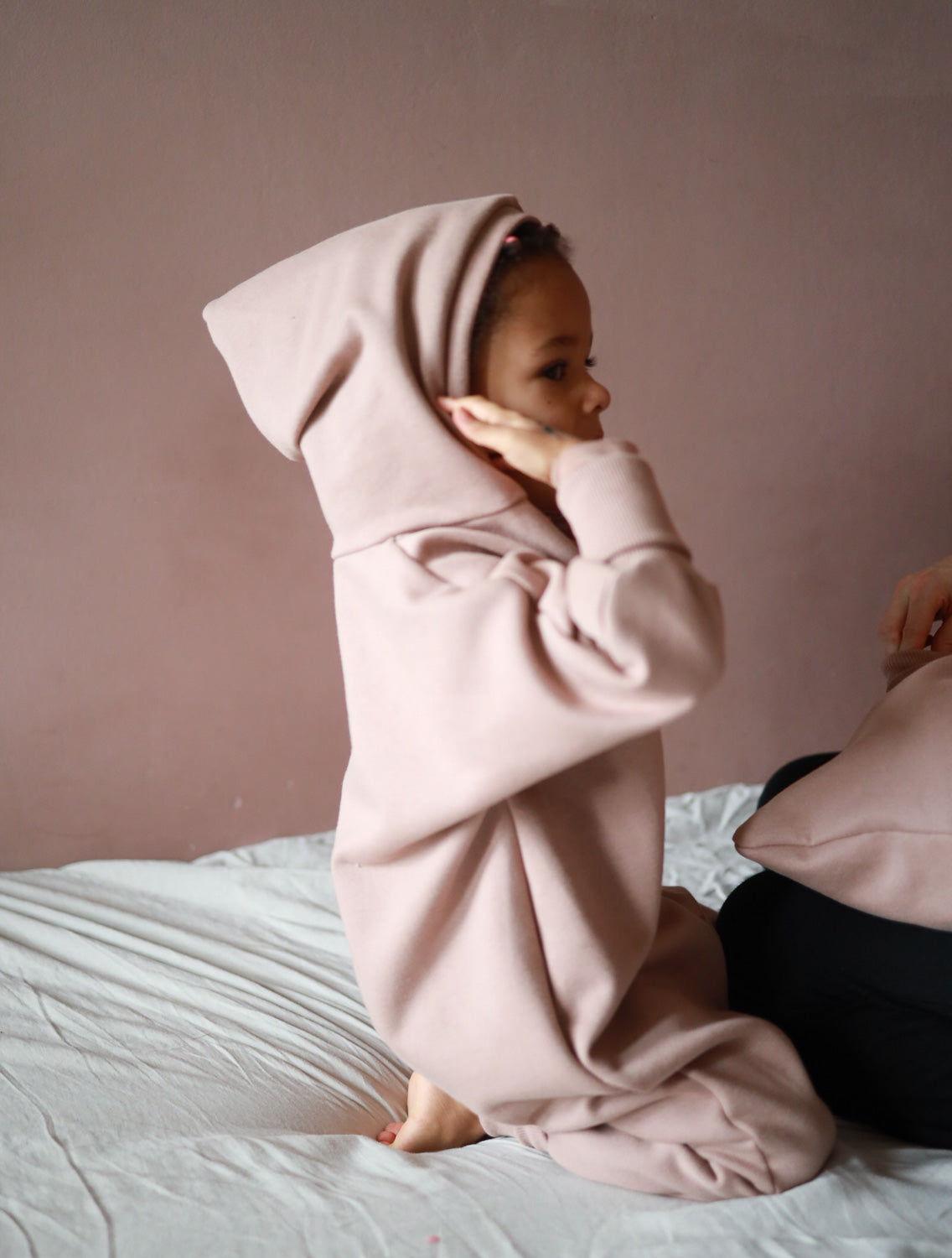 Ein Kind trägt den Kuschelanzug Bio-Sweat - Puder mit Wichtelkapuze, während es auf einem Bett liegt. Der Anzug wirkt weich und gemütlich, ideal für drinnen und draußen.