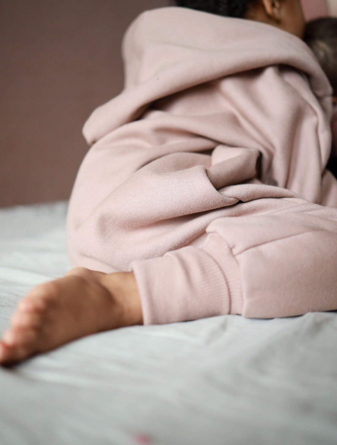 Ein Baby liegt gemütlich auf einem Bett, eingehüllt im Kuschelanzug Bio-Sweat - Puder mit Wichtelkapuze, ideal für Innen- und Außentemperaturen, aus weicher, angerauter Bio-Baumwolle gefertigt.