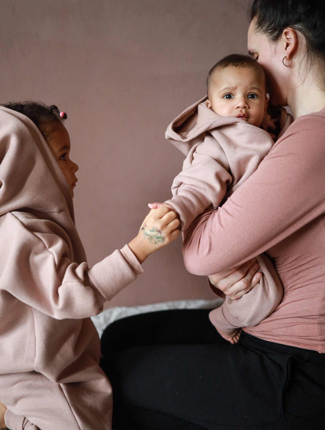 Kuschelanzug Bio-Sweat - Puder: Mutter hält Baby im puderfarbenen Kapuzenanzug, während älteres Geschwisterkind die Hand des Babys ergreift, was eine innige Familienszene zeigt.