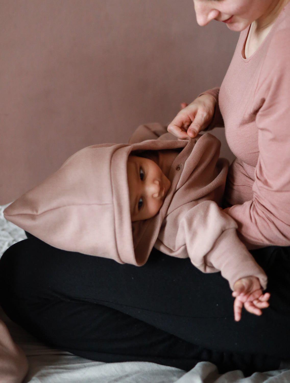 Eine Frau hält ein Baby im Kuschelanzug Bio-Sweat - Puder, einem weichen, kapuzenartigen Einteiler aus Bio-Baumwolle, der gemütlich auf ihrem Schoß ruht, ideal für Komfort und Bewegungsfreiheit.