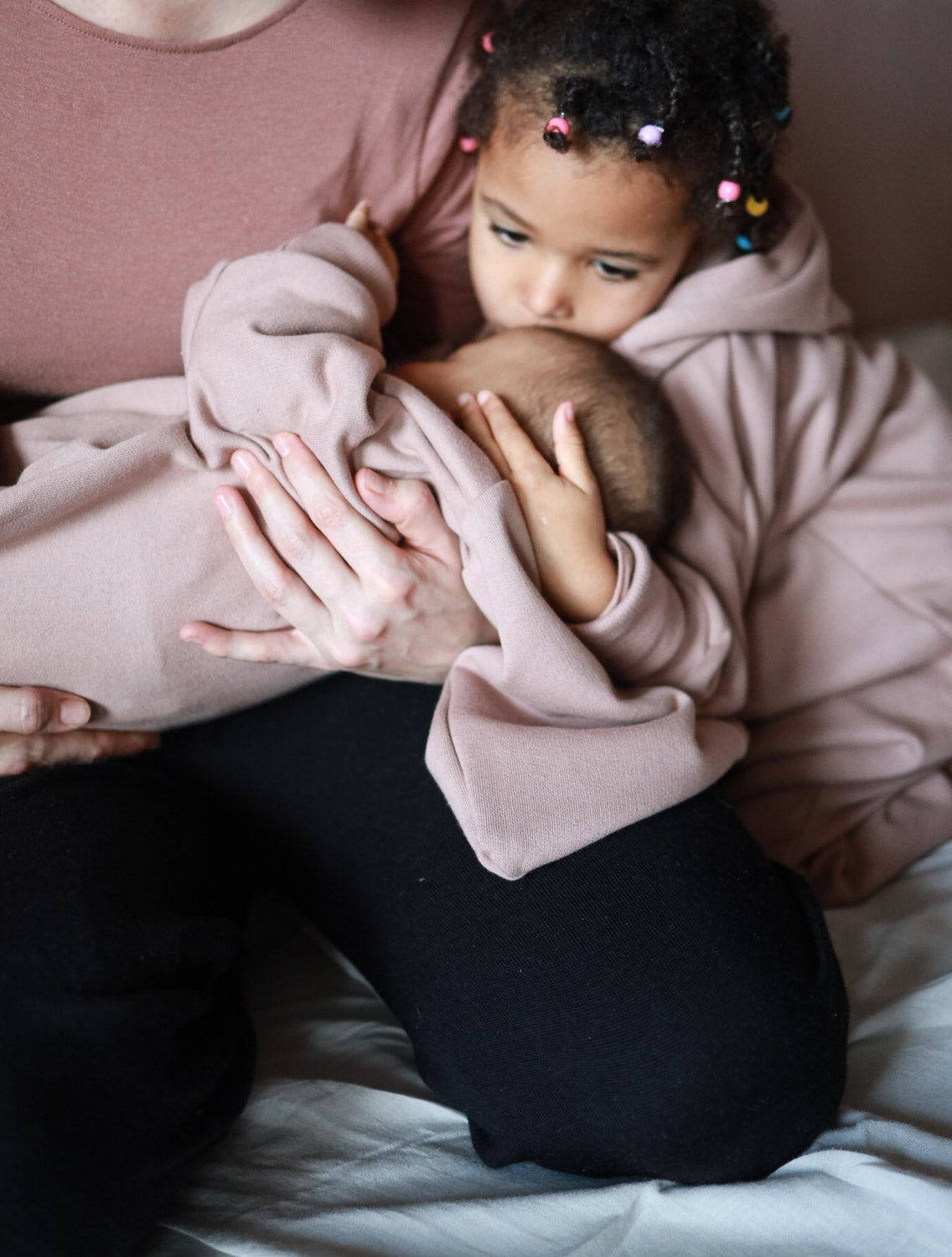 Kind hält Baby mit Kuschelanzug aus Bio-Sweat, Puder. Der Anzug bietet Komfort und Wärme, ideal für drinnen und draußen. Mit Wichtelkapuze, großzügigem Schnitt und Krempelbündchen für mitwachsendes Trageerlebnis.