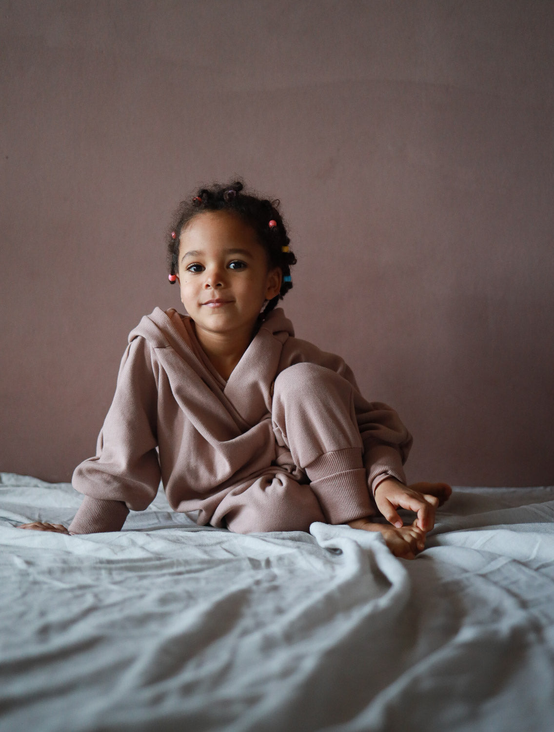 Ein Mädchen sitzt auf einem Bett und trägt den Kuschelanzug Bio-Sweat - Puder mit Wichtelkapuze, ideal für Komfort und Wärme aus Bio-Baumwolle.