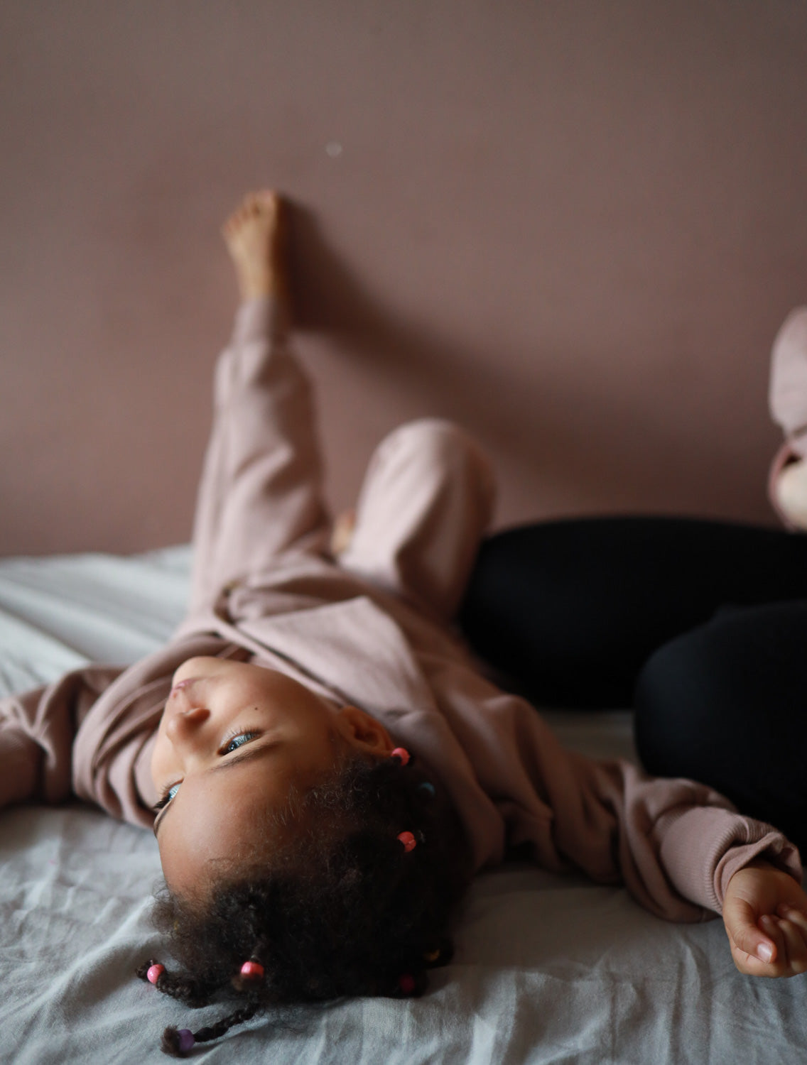 Ein Kind liegt auf einem Bett, trägt einen Kuschelanzug Bio-Sweat - Puder mit kleinen Zöpfen und rosa Perlen, neben Erwachsenbeinen in schwarzen Leggings, auf einem hellgrauen Bettlaken.