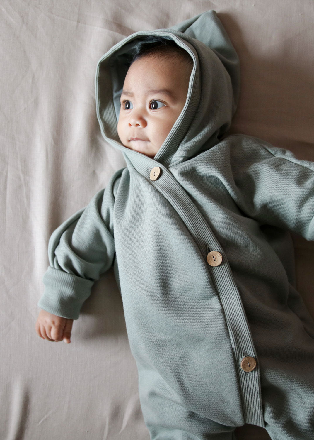 Kuschelanzug Bio-Sweat - Aqua: Baby liegt auf dem Bett, trägt einen kuscheligen Overall mit Wichtelkapuze, sichtbar weiche, angeraute Sweat-Bio-Baumwolle, großzügiger Schnitt, Krempelbündchen an Armen und Beinen.  Erstausstattung Baby und Baby Geschenk