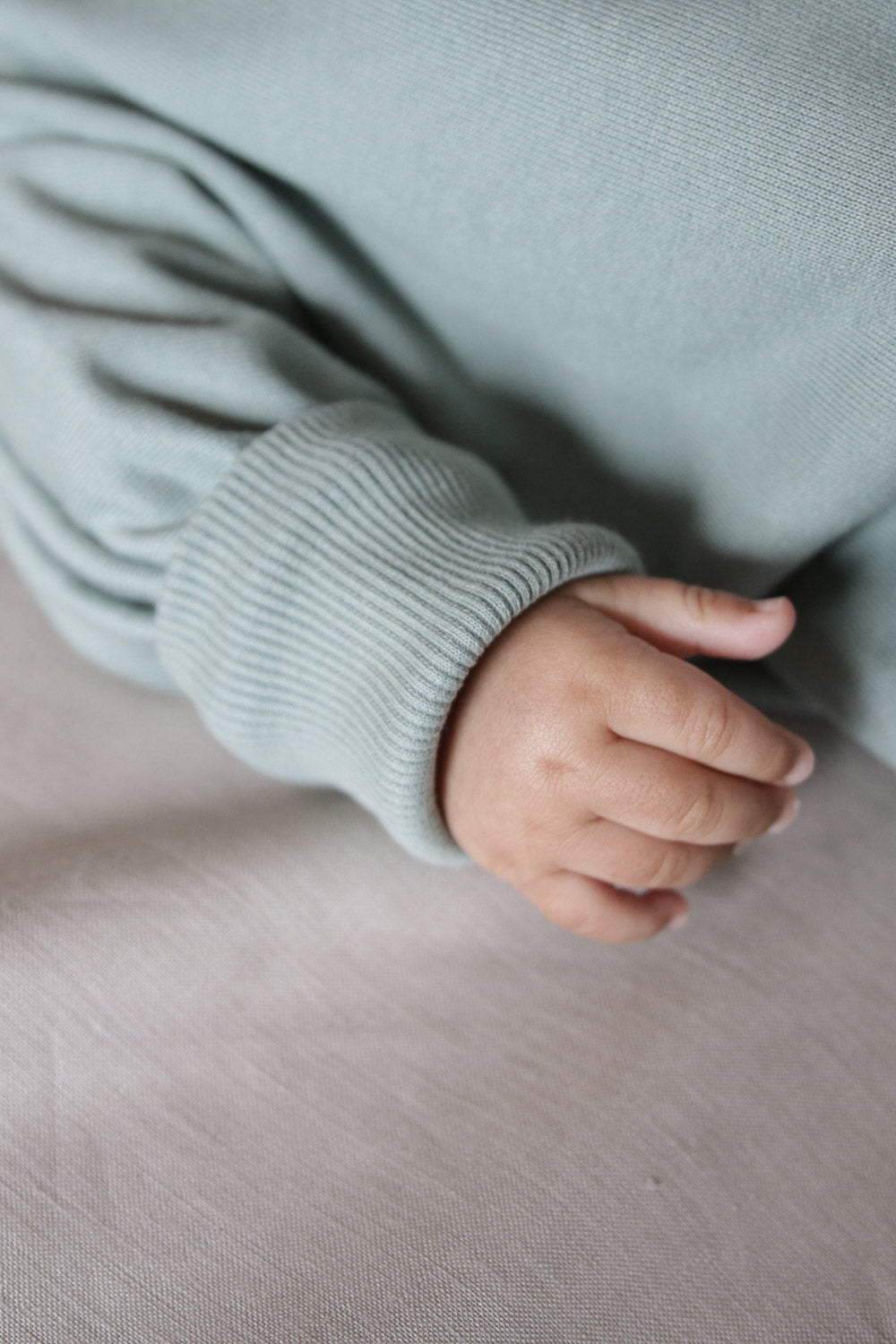 Kuschelanzug Bio-Sweat - Aqua: Nahaufnahme einer Babyhand, die sanft auf weichem, weißem Stoff ruht, der den kuscheligen Overall aus Bio-Baumwolle darstellt, ideal für Komfort und Wärme.  Erstausstattung Baby und Baby Geschenk zur Geburt.