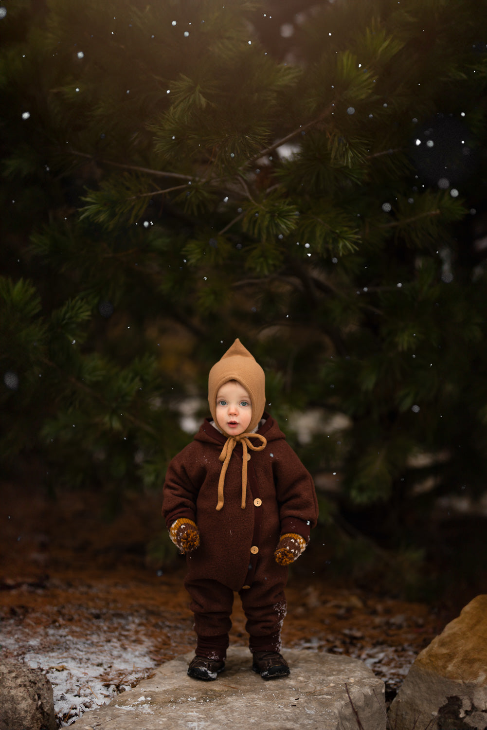 Ein Kind trägt den Kuschelanzug Wollwalk - Kastanie aus 100% Schurwolle im Schnee, ideal für kühle Jahreszeiten, mit wärmenden und atmungsaktiven Eigenschaften.