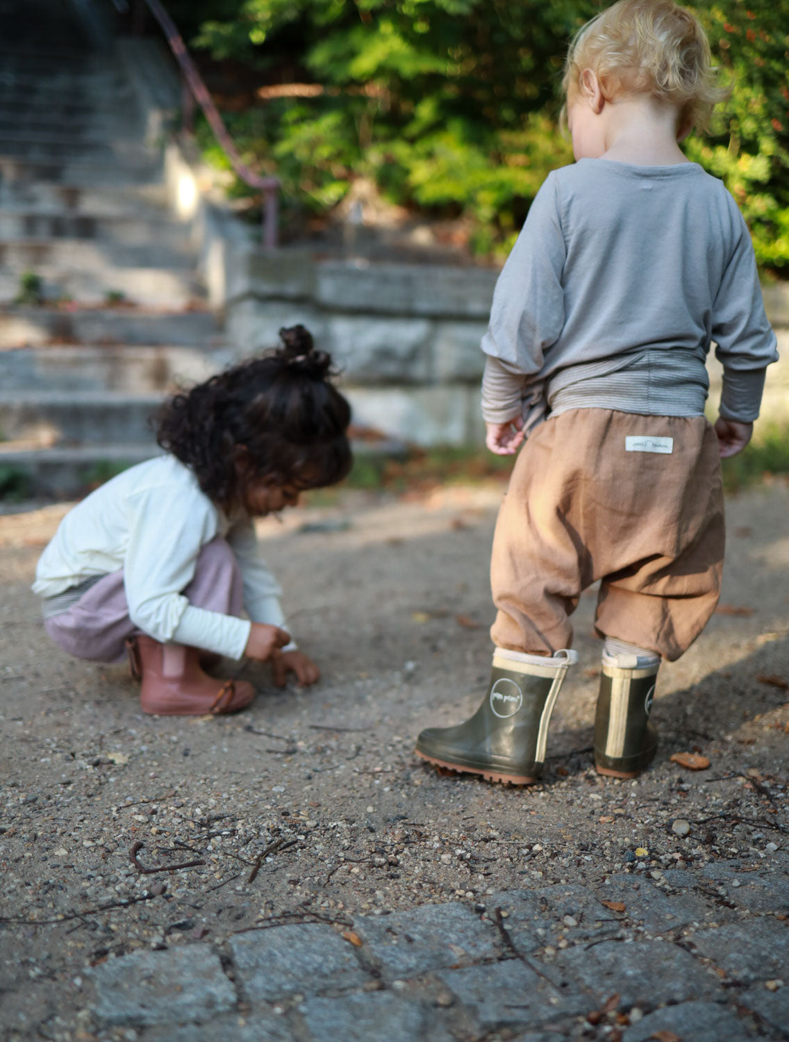 Zwei Kinder spielen draußen; eines trägt die Knickerbocker Leinen - Weizen, eine luftige, bequeme Hose mit elastischen Bündchen, ideal für Sommer und Winter, gefertigt aus robustem, atmungsaktivem Leinen.