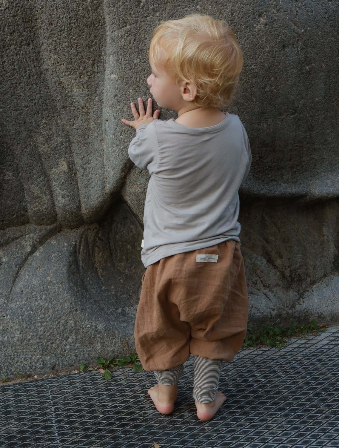 Kind berührt einen Felsen, trägt Knickerbocker Leinen - Weizen, bequeme, elastische Hose aus robustem, gewaschenem Leinen mit weitem Schnitt und Kängurutasche, ideal für warme und kühle Tage.