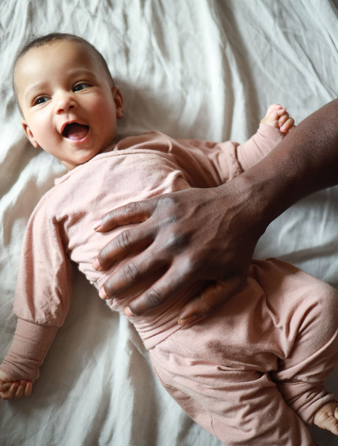Baby in einem Loopbody Kaschmir-Mix - Puder liegt auf einem Bett. Der Body umschließt den Bauch, ohne Knöpfe, für einfaches Anziehen, aus weichem, atmungsaktivem Material designed.  Erstausstattung Baby und Baby Geschenk zur Geburt.