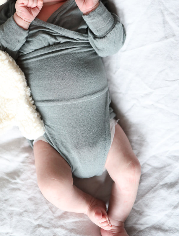 Ein Baby trägt den Loopbody Kaschmir-Mix von PETIT COCHON Berlin, der ohne Knöpfe und mit einem weichen, anschmiegsamen Design auf einem Bett liegt.