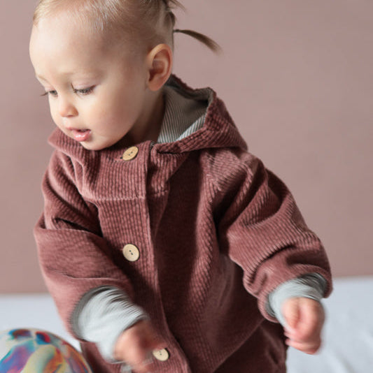 Ein Baby trägt die Wichteljacke Breitcord - Rosenholz mit süßer Wichtelkapuze, spielt mit einem Ball und hält einen Luftballon.