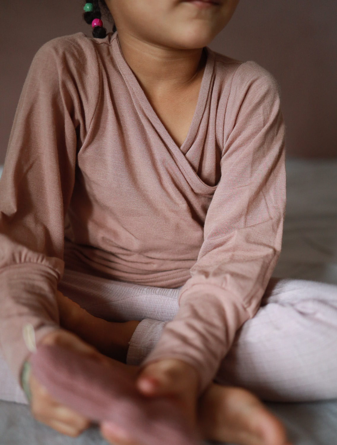 Person sitzt auf Bett, trägt Wickelshirt aus Kaschmir-Mix - Puder, das weich und elastisch ist, ideal als Shirt oder Unterhemd für Kinder.