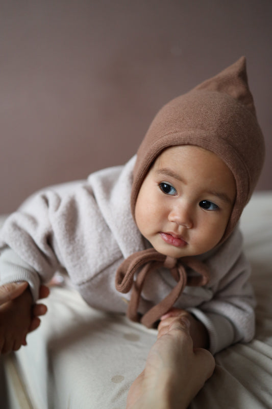 Baby trägt die Zwergenmütze Strick - Nougat, kuschelig weich aus recycelter Baumwolle mit Bindeband, perfekt passend für Herbst- und Winteroutfits. Erstausstattung Babys. Perfekte Baby Geschenke.