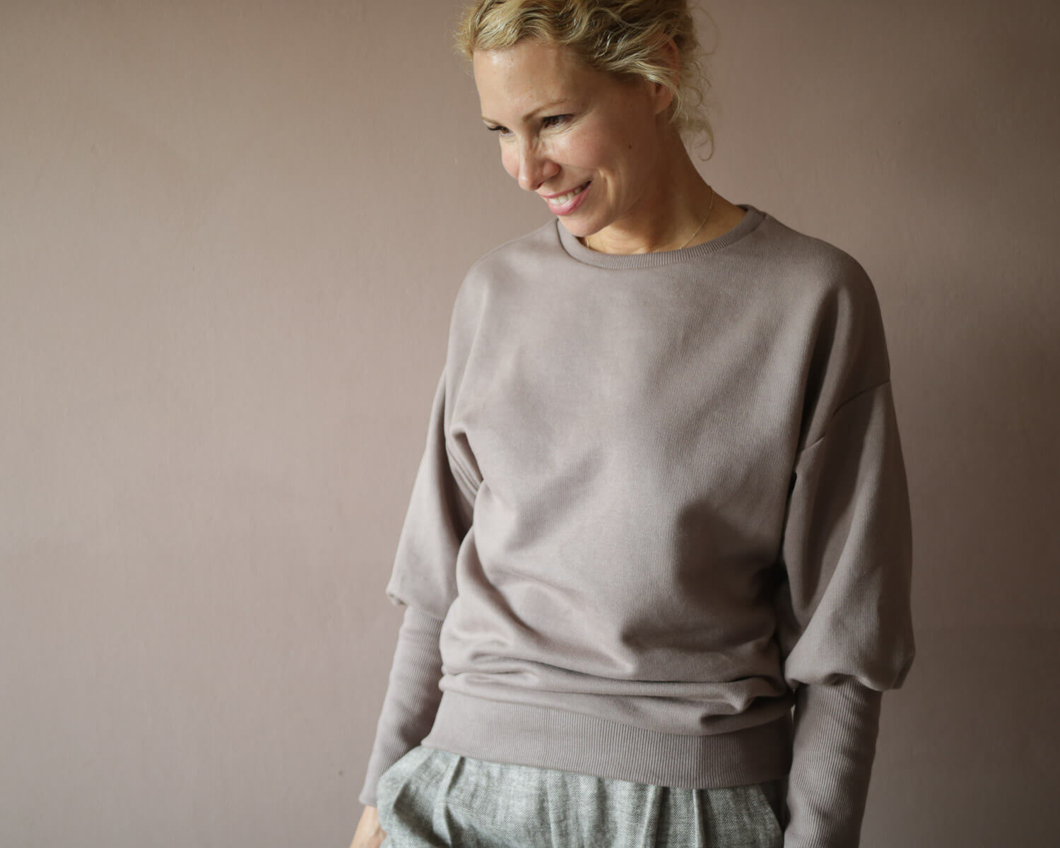 Eine lächelnde Frau in einem grauen Pullover von Petit Cochon,  Nahaufnahme des Gesichts und des Stoffes. Sie trägt auch eine Bundfaltenhose Fischgrat.