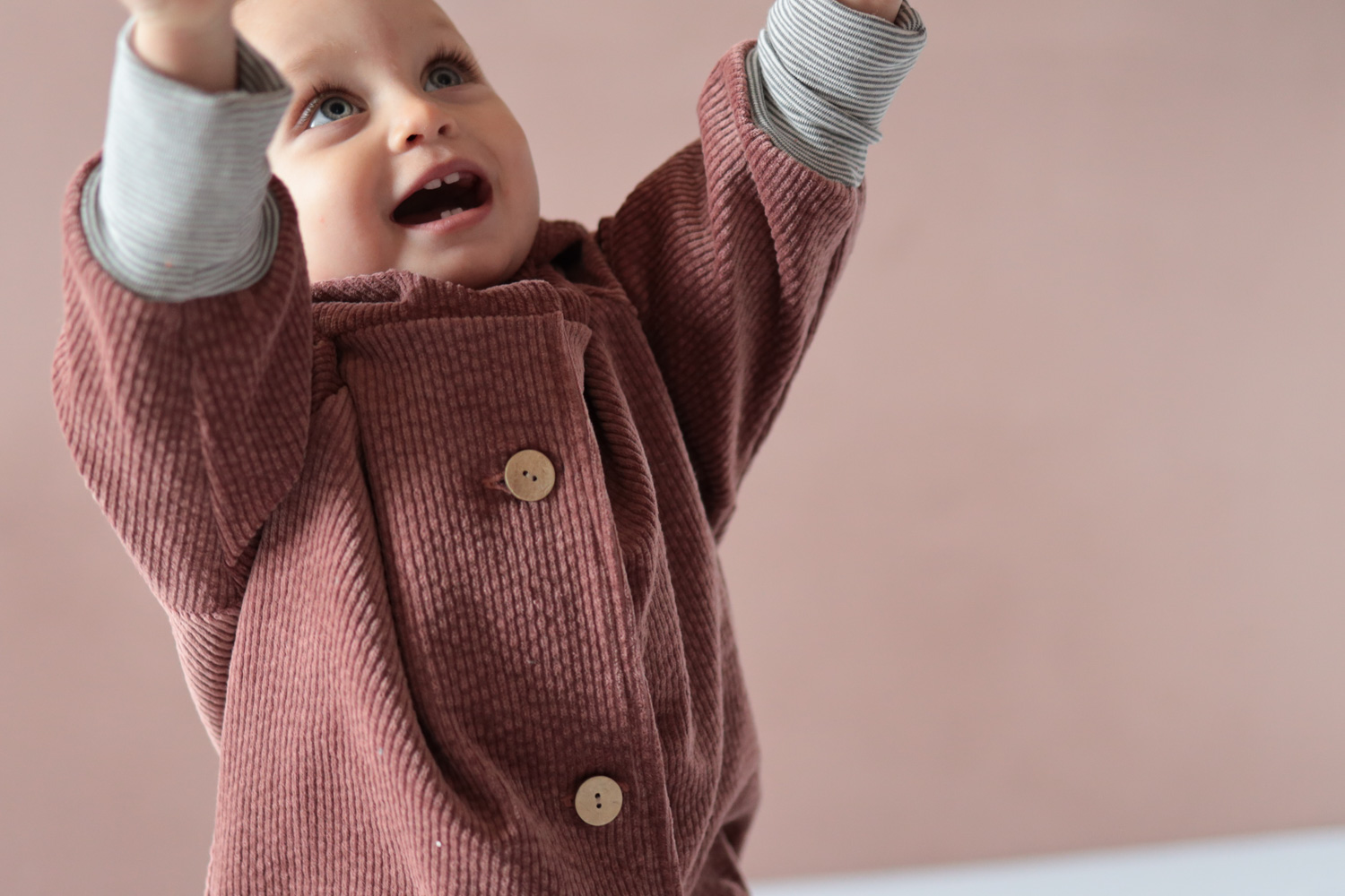 Ein Kind in eine Wichteljacke Breitcord Rosenholz  mit erhobenen Armen und überrascht wirkendem Gesichtsausdruck, Nahaufnahme von Knöpfen und Ärmeln mit Ringelbündchen sichtbar. Nachhaltige Kinderkleidung von Petit Cochon.