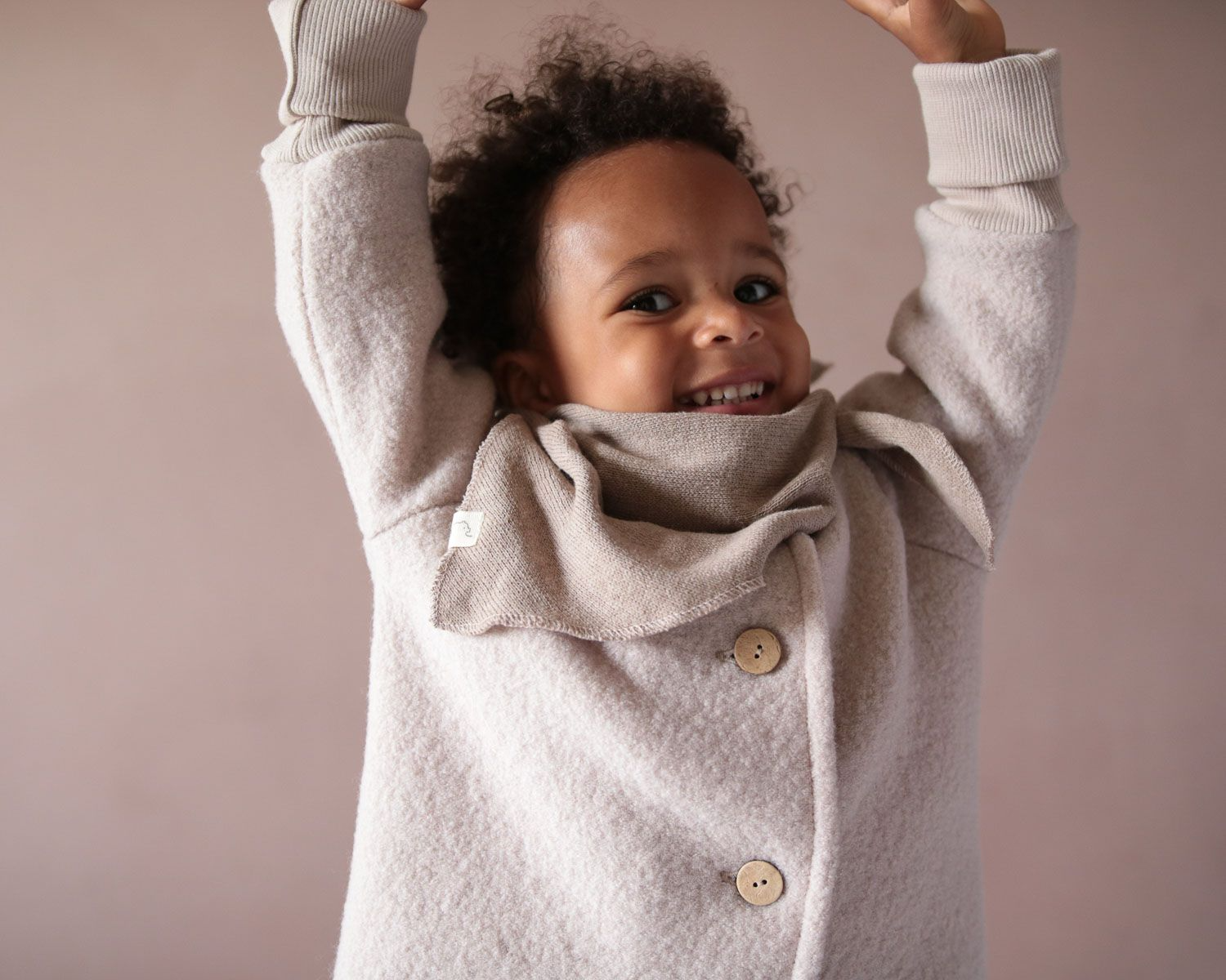 Ein lächelndes Kleinkind mit lockigem Haar, trägt eine Wollwalk Jacke für Kinder und ein Stricktuch von Petit Cochon.
