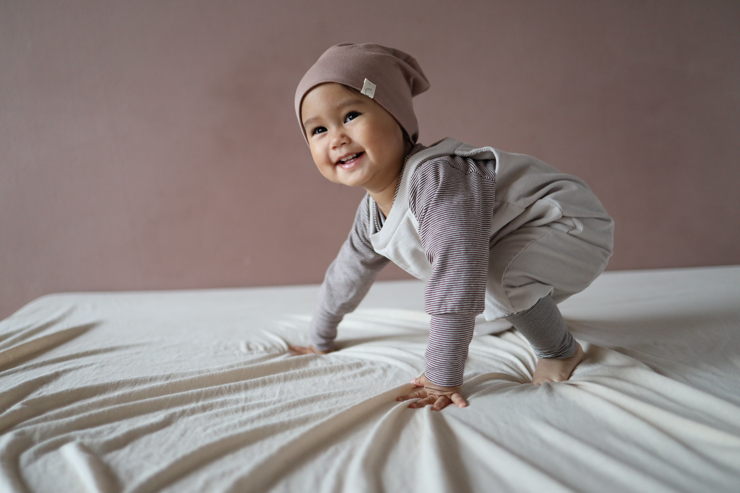 Ein lächelndes Baby mit Beanie, Latzhose und Loopbody der Baby Body, krabbelt auf einem Bett, 