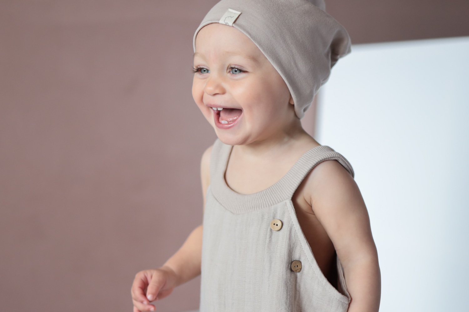 Ein lächelndes Baby trägt eine Beanie und eine Schlupflatzhose von Petit Cochon, mit einem detaillierten Fokus auf das Gesicht und die Kleidung.