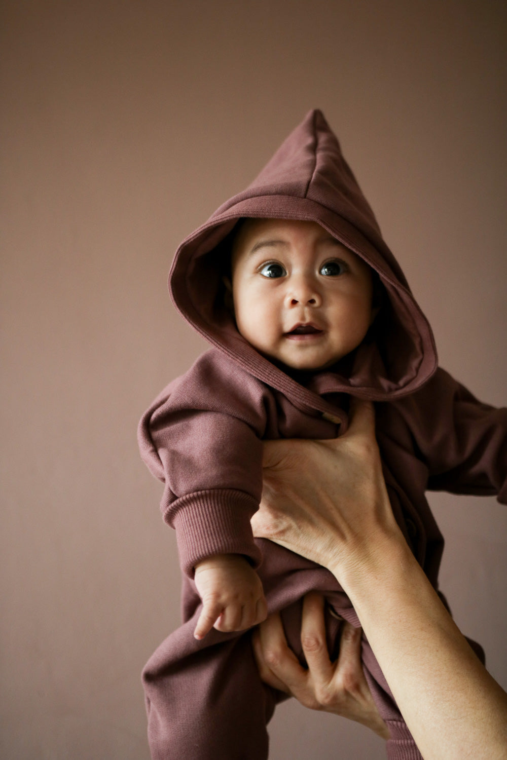 Baby mit Overall aus Bio-Baumwolle in lilabraun , gehalten von einem Erwachsenen.
